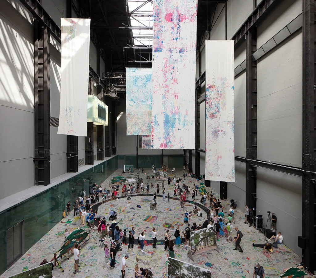 Inštalačný pohľad na dielo Eia Arakawu-Nasha Mega Please Draw Freely v Tate Modern v Londýne, 2021 — veľké maľované plátna visiace zo stropu, návštevníci kreslia na podlahu.