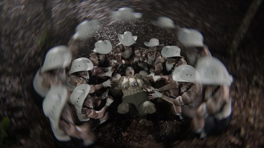 Venice Biennale 2026 — Natasha Tontey, The Phantom Combatants, 2026, video still, postavy v bielych klobúkoch obklopujú postavu na zemi, fisheye objektív.
