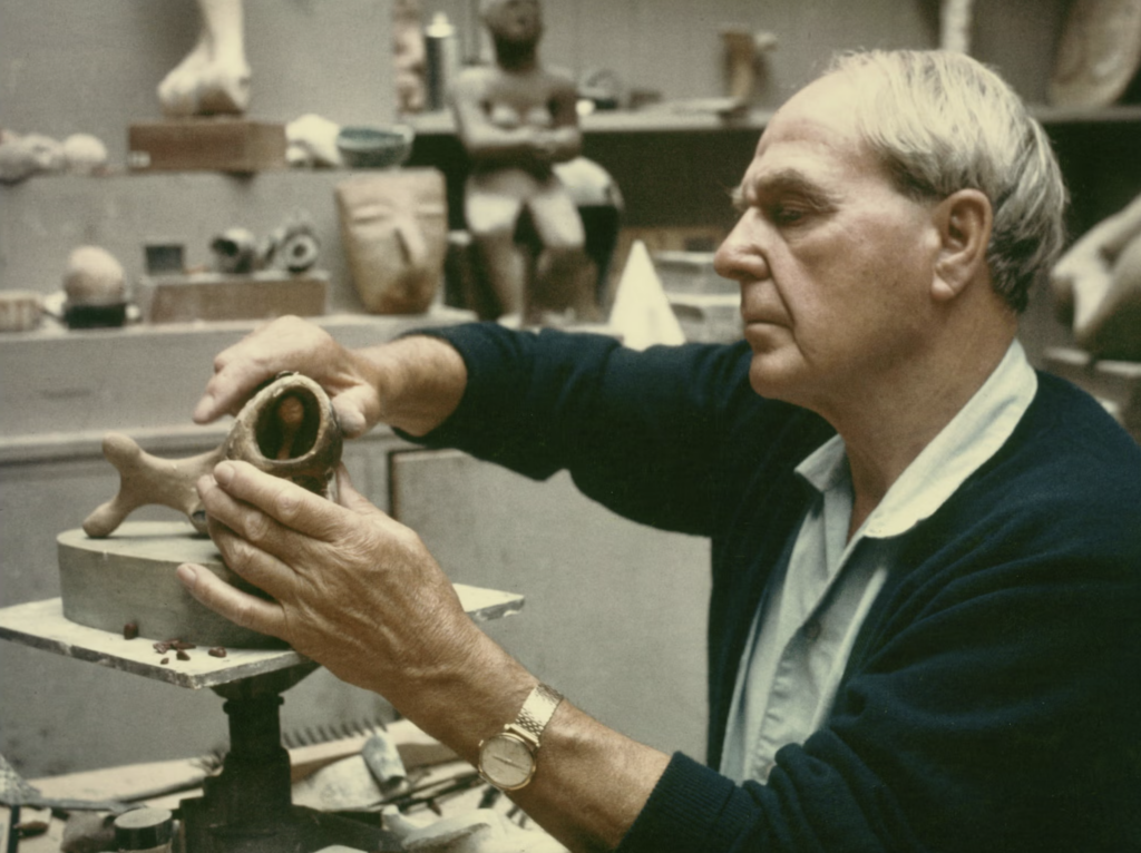 Henry Moore pracuje na sadrovej maketo vo svojom ateliéri okolo roku 1968, v pozadí socha Seated Figure z roku 1929 a hlava v aztéckom štýle na poličke
