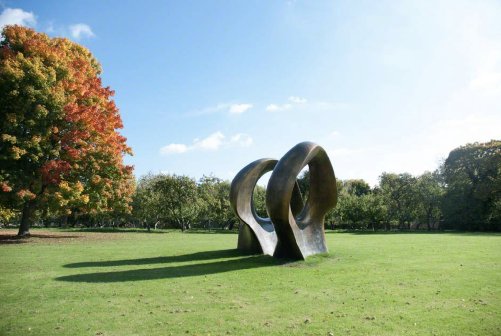 Bronzová socha Double Oval od Henryho Moora na lúke pri Henry Moore Studios and Gardens, v pozadí strom s jesenným lístím
