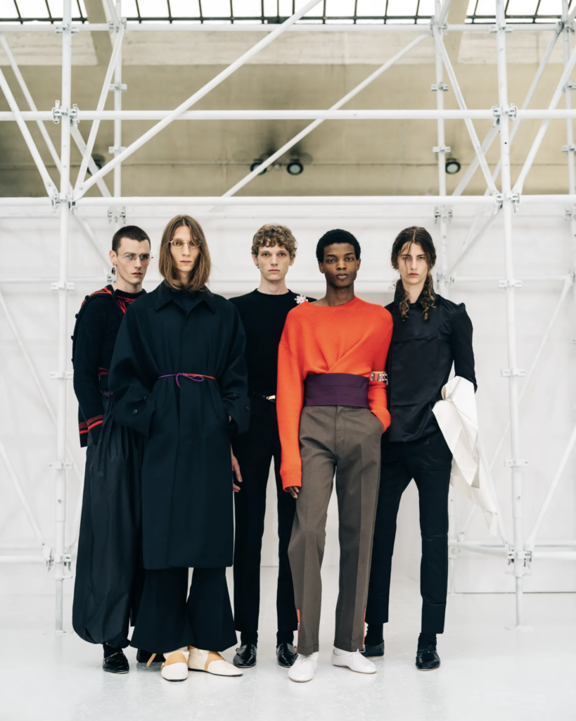 Päť modelov backstage kolekcie Celine Heat stojí pred konštrukciou lešenia, v outfitoch v čiernej, oranžovej a olivovej farbe