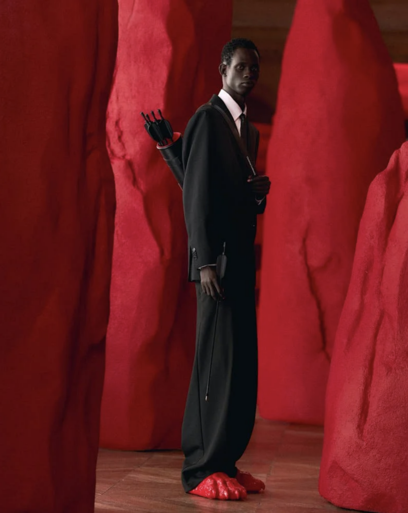 Model v čiernom plášti stojí medzi červenými organickými sochárskymi formami počas prezentácie Christian Louboutin Men SS27, na nohách červené sandále Claw Slide