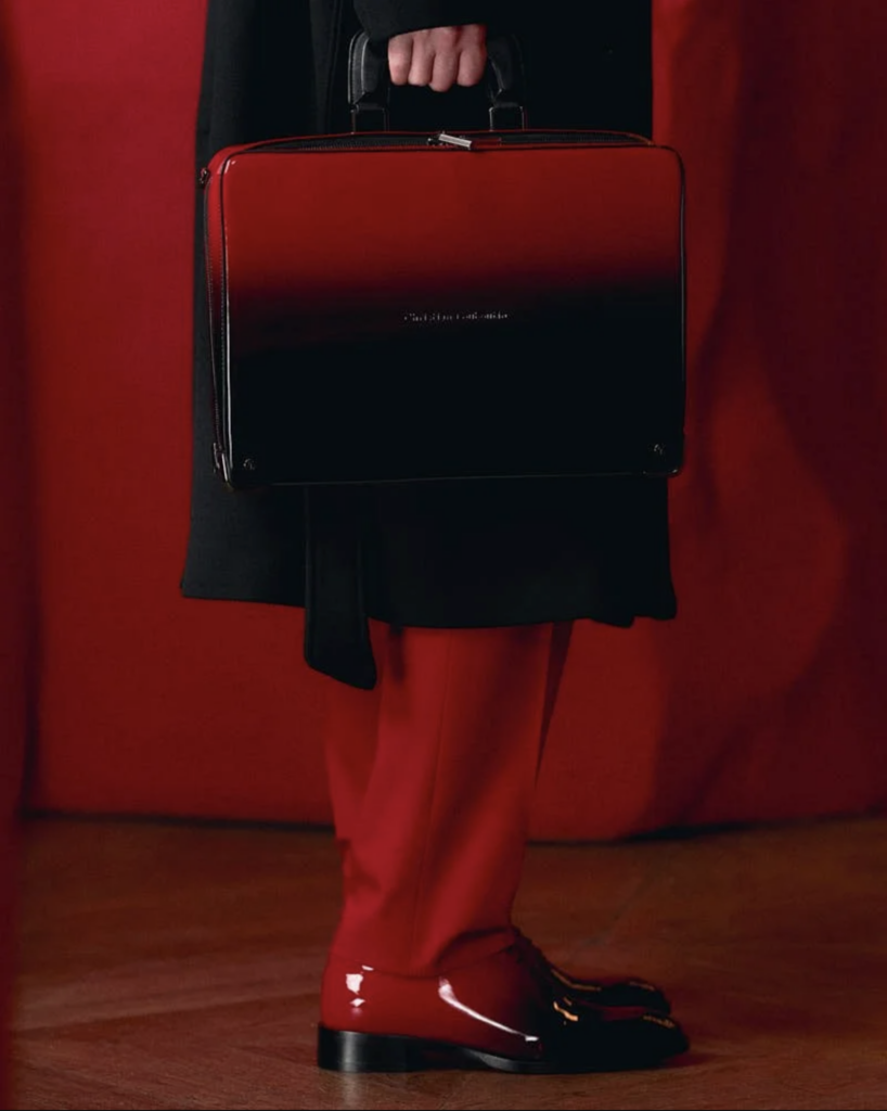 Detail nôh modela v čiernych lakovaných mokasínoch s červenými ponožkami a čiernou kabelkou pri prezentácii Christian Louboutin Men SS27
