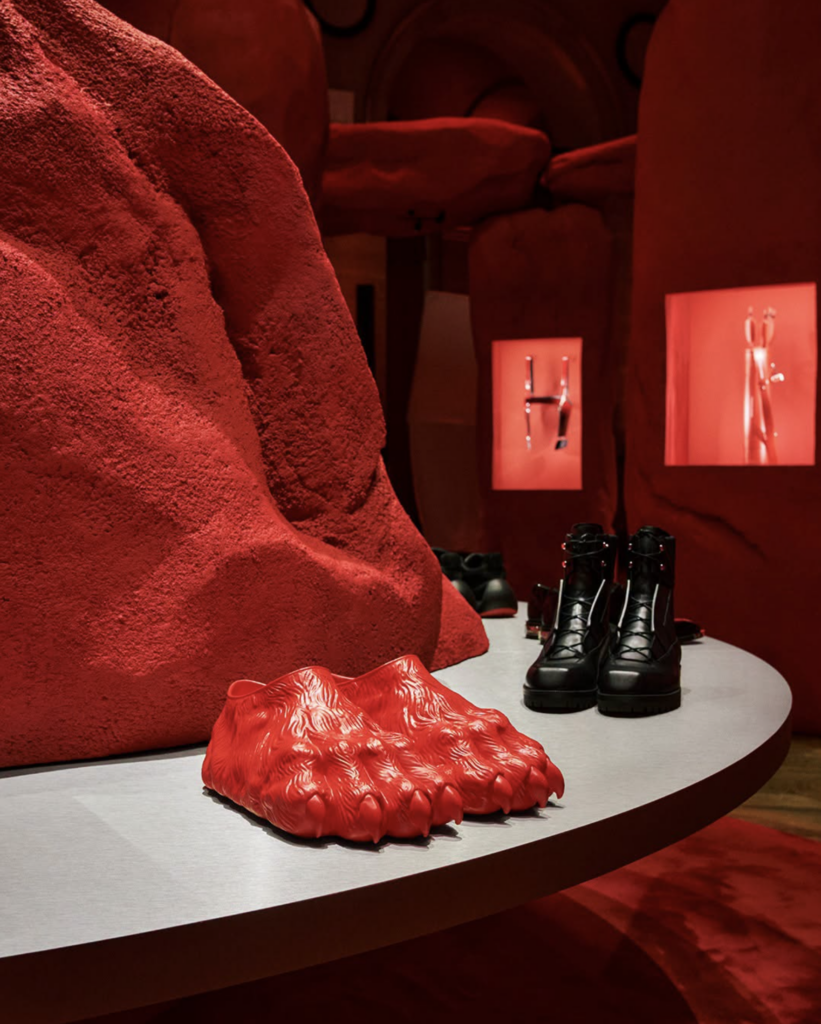Detail červených sandálov Claw Slide v tvare zvieracieho pazúra vedľa čiernych šnurovacích čižiem na podstavci v rámci prezentácie Christian Louboutin Men SS27