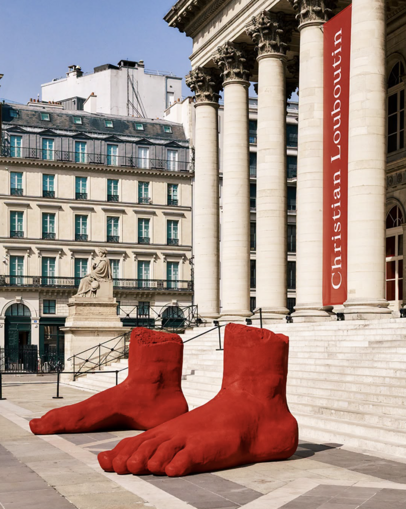 Dve monumentálne červené sochárske nohy pred stĺporadím Palais Brogniart v Paríži s červenou zástavou Christian Louboutin
