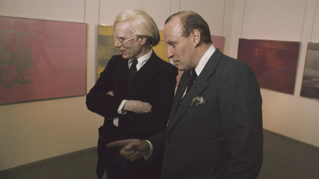 Andy Warhol a Bruno Bischofberger spolu v galerijnom priestore s farebnými obrazmi v pozadí, čiernobiela archívna fotografia