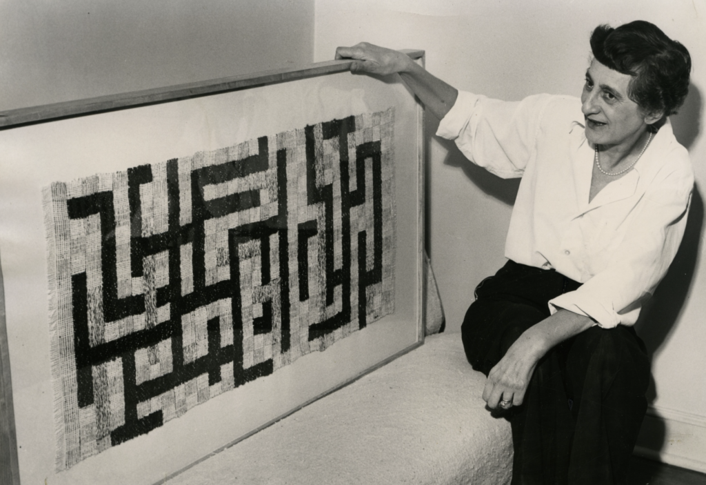 Čiernobiela archívna fotografia Anni Albers pri práci na tkanom diele, umelkyňa drží drevený rám a pozerá sa na textilnú kompozíciu