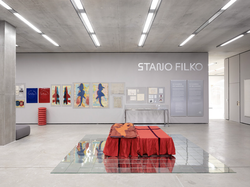 Inštalačný pohľad na výstavu Stana Filka 12 Chakras of Becoming v Museum der Moderne Salzburg s farebnými grafikami na stene a červeným objektom na sklenej podlahe v popredí