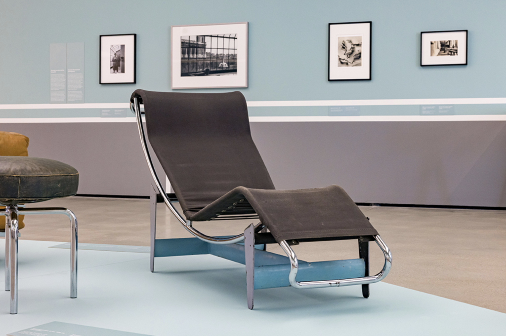 Inštalačný pohľad na výstavu Charlotte Perriand v Museum der Moderne Salzburg s dreveným nábytkom na ružovej podlahe a veľkoformátovou čiernobielou fotografiou interiéru v pozadí