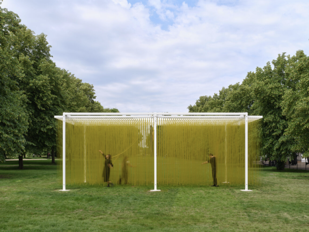 Traja návštevníci prechádzajú cez žlté PVC rúrky sochy Pénétrable BBL Jaune od Jesúsa Rafaela Sota na trávniku v Kensington Gardens, ich siluety sú rozmazané pohybom