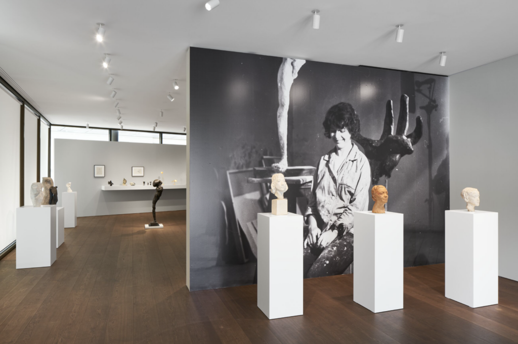 Inštalačný pohľad na výstavu Aliny Szapocznikow v Hauser & Wirth Zürich, malé sochárske hlavy na bielych podstavcoch pred veľkoformátovou čiernobielou fotografiou umelkyne v ateliéri