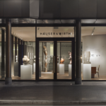 Presklený interiér pobočky Hauser & Wirth na Bahnhofstrasse v Zürichu, žena prechádza popri veľkých sochách, medzi nimi hnedej diskovej soche a bielej svetelnej inštalácii