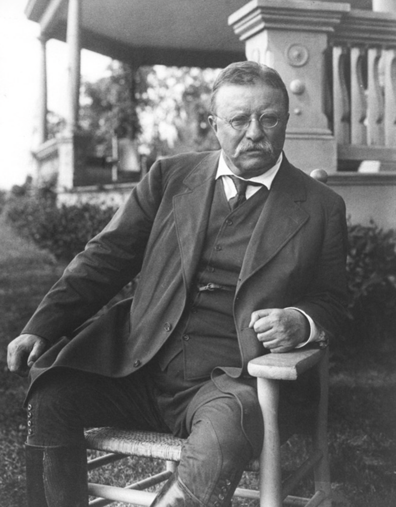 Čiernobiela fotografia Theodora Roosevelta sediaceho na verande jeho domu Sagamore Hill, oblečeného v obleku, s okuliarmi a fúzmi