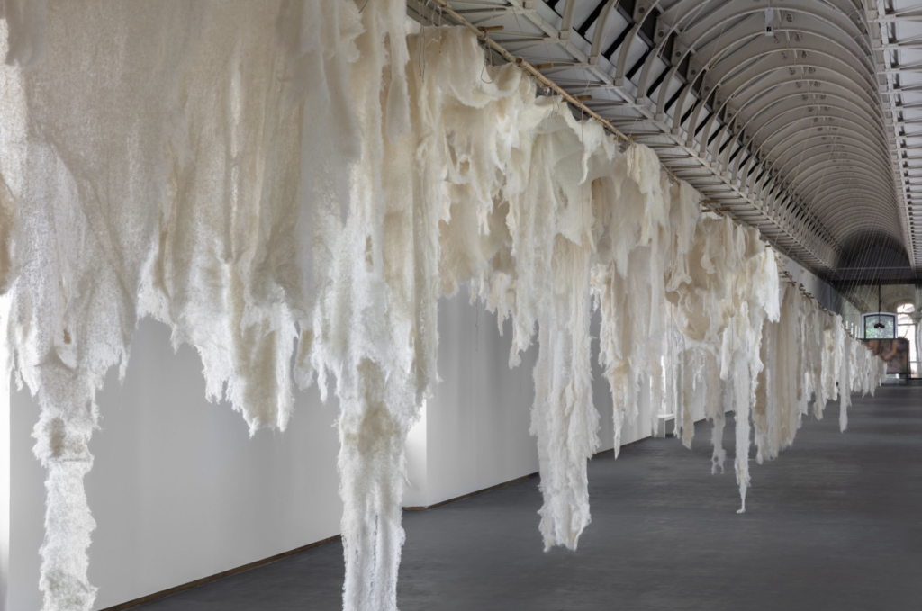 Alt text:
Inštalačný pohľad na výstavu Cecilie Vicuñe El glaciar ido v Castello di Rivoli, monumentálne biele quipu visiace v presvetlenej šachte historickej budovy