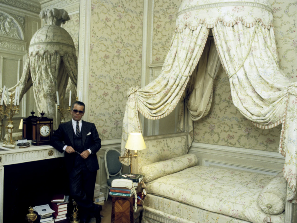 Karl Lagerfeld stojaci pri krbe vo svojej spálni vo Villa La Vigie na Azúrovom pobreží, v pozadí baldachýnová posteľ s kvetovaným textilom