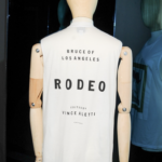 Drevená figurína oblečená v bielom tričku s nápisom "Bruce of Los Angeles Rodeo, edited by Vince Aletti"