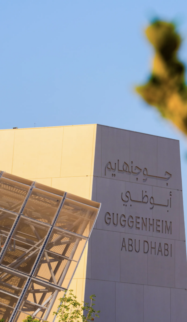 Detail vstupného pilóna Guggenheim Abu Dhabi s nápisom v arabčine a angličtine vedľa kovovej a sklenenej konštrukcie budovy