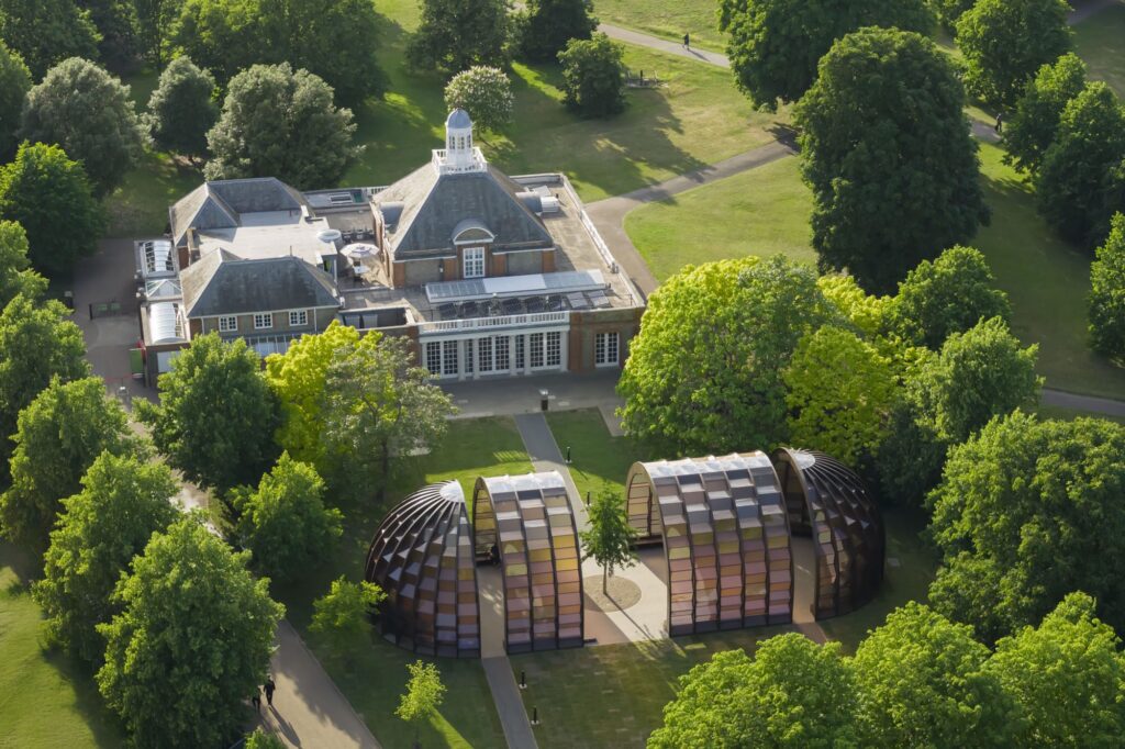 Letecký pohľad na pavilón Serpentine 2025 A Capsule in Time od Mariny Tabassum, drevené oblúkové konštrukcie s farebnými panelmi medzi stromami v Kensington Gardens