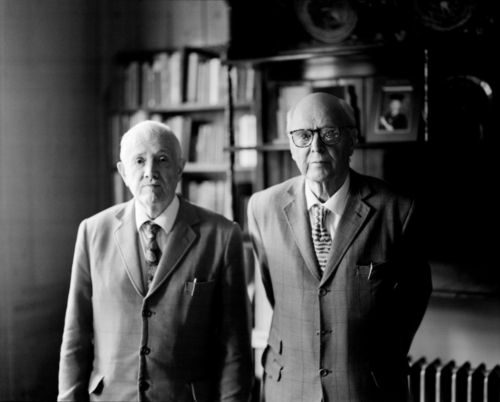 Čiernobiely portrét dvojice Gilbert & George v obleku, stojacich vedľa seba v knižnici