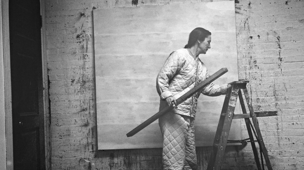 Čiernobiela archívna fotografia Agnes Martin stojacej na rebríku v ateliéri, maľujúcej veľké štvorcové plátno, oblečenej v prešívanom pracovnom overale.