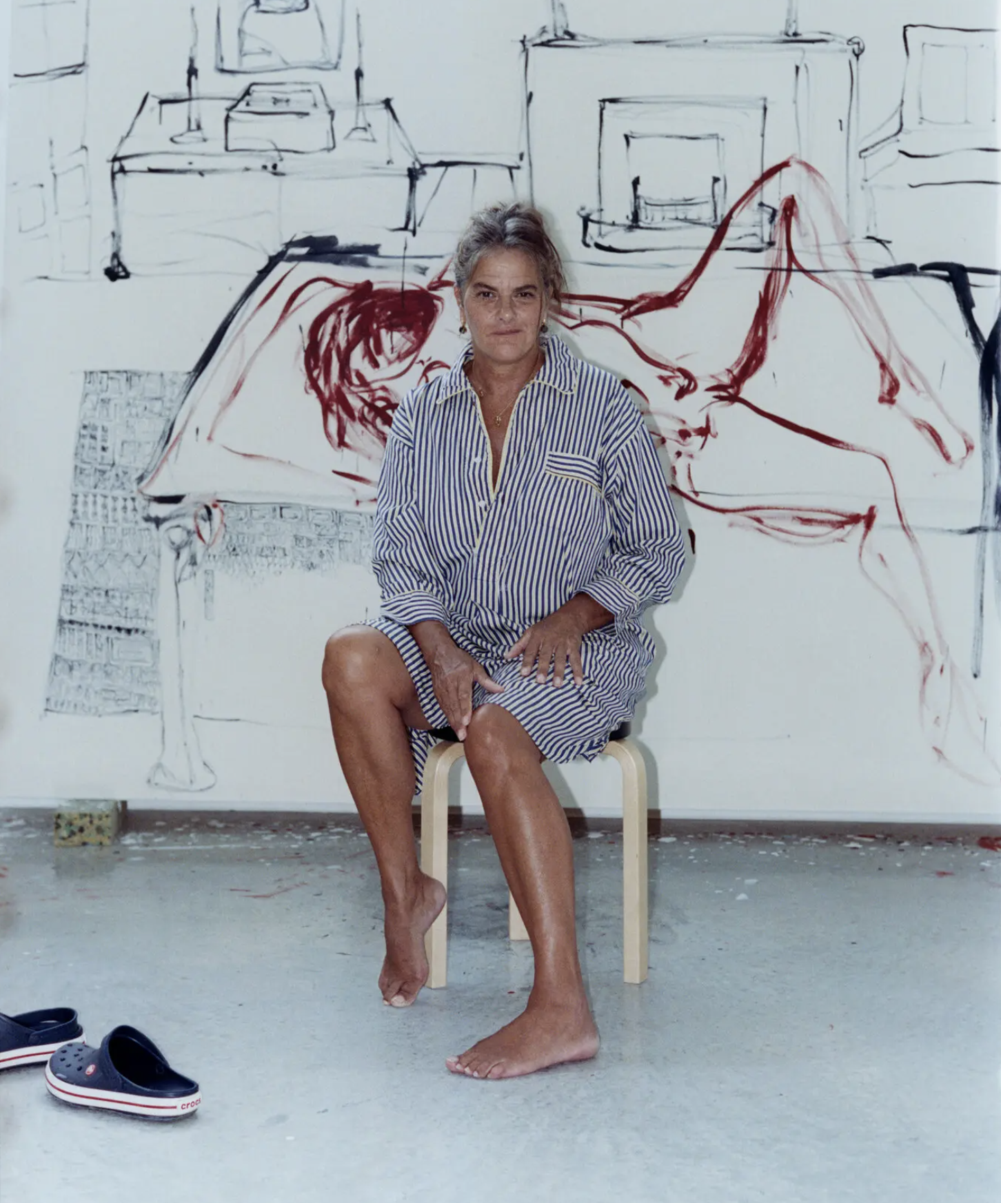 Tracey Emin sedí na drevenej stoličke vo svojom ateliéri v Margate, bosá, oblečená v pruhovanej košeli, za ňou nedokončený obraz s expresívnymi ťahmi štetca v čiernej a červenej farbe.