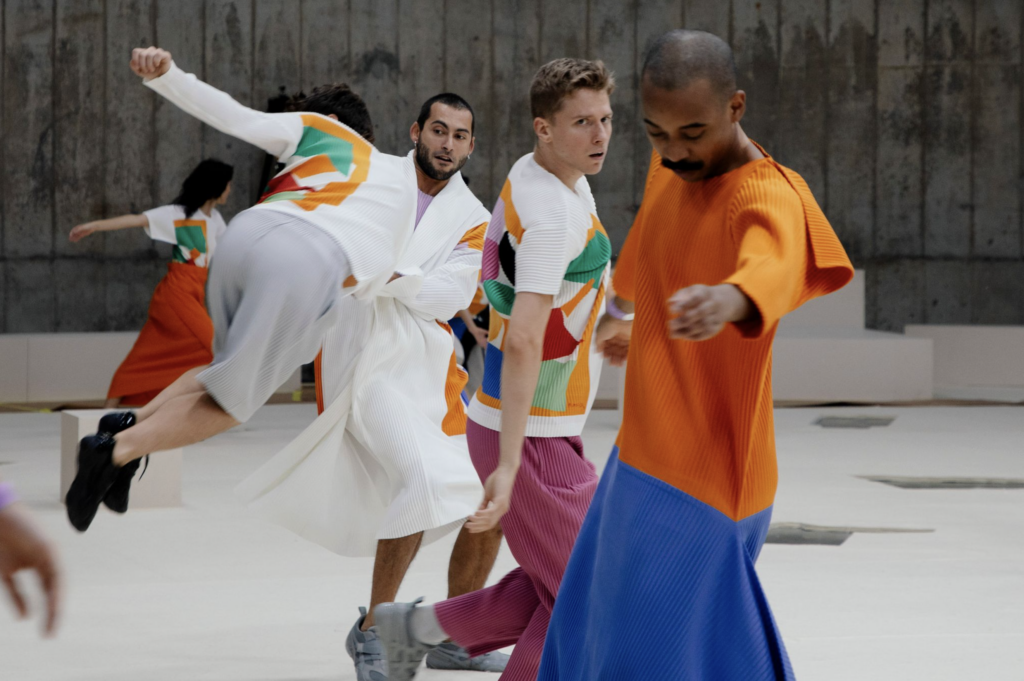 Tanečníci Dresden Frankfurt Dance Company v pohybe, oblečení v plisovaných kúskoch HOMME PLISSÉ ISSEY MIYAKE s farebnými geometrickými vzormi inšpirovanými Matissom, jeden tanečník uprostred skoku.